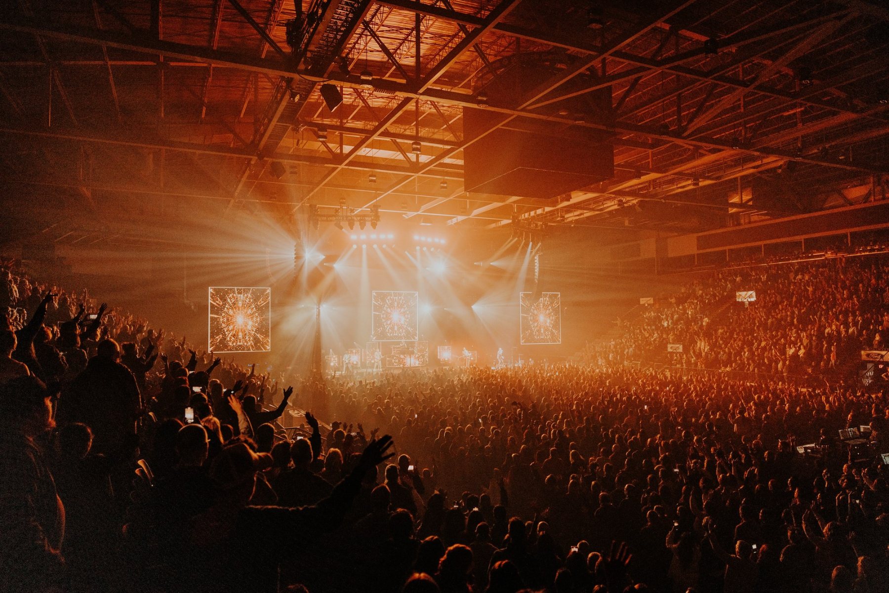Packed arena under a steel roof, stage lights cutting through haze over a crowd with raised hands